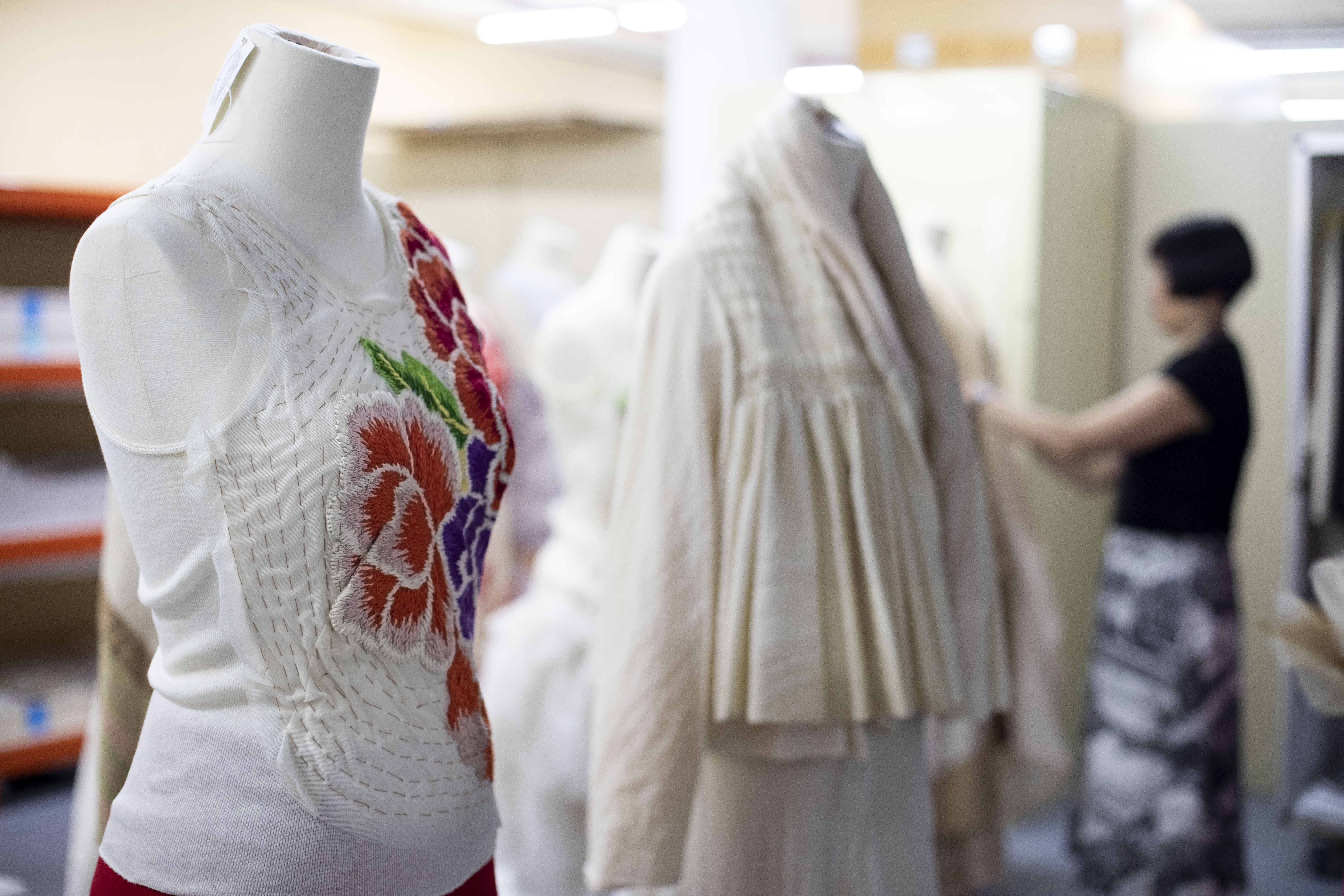 A white sleeveless top with floral applique pattern on a mannequin. In the background, out of focus, are a second dressed mannequin, and beyond that, a woman dressing a third mannequin.